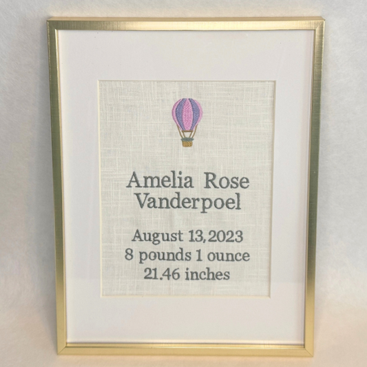 Framed embroidery with a hot air balloon illustration and baby details on a light background
